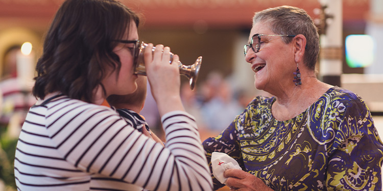 Young female member drinks from a chalice; another female member smiles and pats her shoulder.